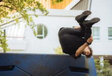Person performing a parkour flip over a blue urban structure amidst cityscape.