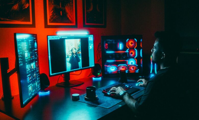A young man working on a multi-screen computer setup in a dark, neon-lit room.
