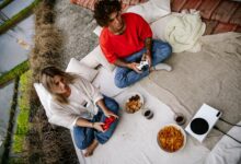 A couple outdoors playing video games with wireless controllers, snacks, and drinks.