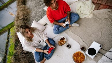 A couple outdoors playing video games with wireless controllers, snacks, and drinks.