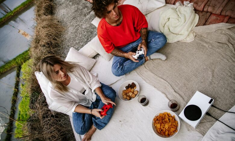 A couple outdoors playing video games with wireless controllers, snacks, and drinks.