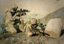 Two soldiers in camouflage uniforms practice tactical maneuvers in a desert environment, exhibiting teamwork and precision.