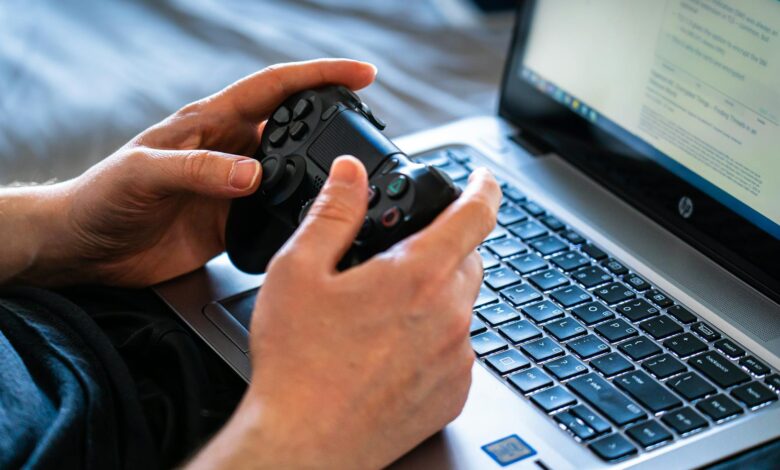 Person holds a gaming controller while interacting with a laptop, emphasizing modern gaming technology.