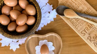 A top-down view of brown eggs in a wooden bowl, with a heart-shaped dish and utensils on a rustic table.