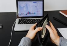 Man holding a leather wallet with bills in front of a laptop displaying financial graphs.