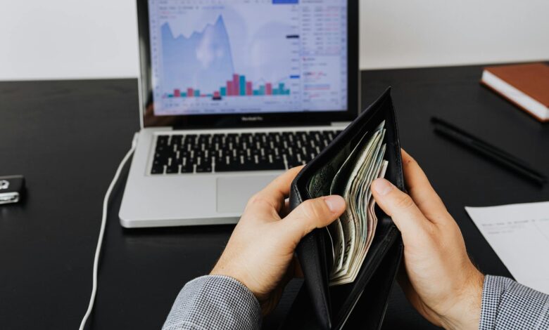 Man holding a leather wallet with bills in front of a laptop displaying financial graphs.