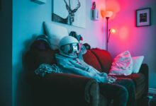 Anonymous guy in VR helmet sitting on sofa with controller while playing video game in cozy room with dark light