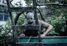 A spooky masked figure peers through a ladder in an eerie overgrown garden setting.