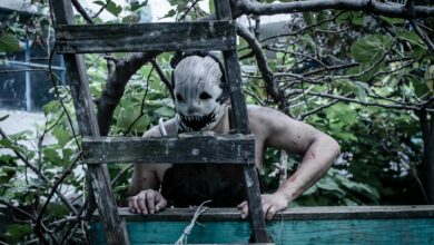 A spooky masked figure peers through a ladder in an eerie overgrown garden setting.