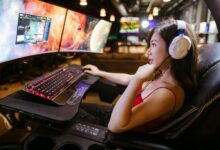 Young woman in red top gaming on dual monitors with white headphones in a modern setup.
