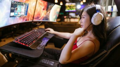 Young woman in red top gaming on dual monitors with white headphones in a modern setup.
