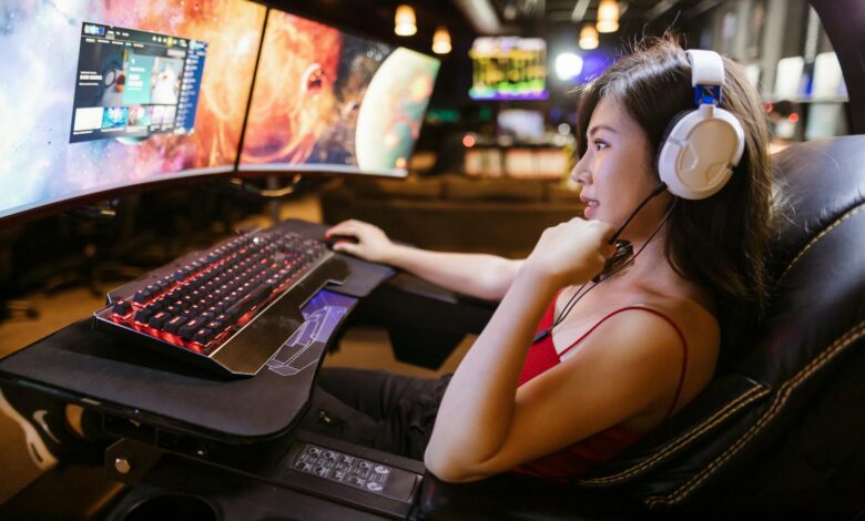 Young woman in red top gaming on dual monitors with white headphones in a modern setup.