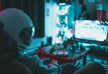 Side view of unrecognizable person in virtual reality helmet sitting on sofa and playing with gamepad in dark room