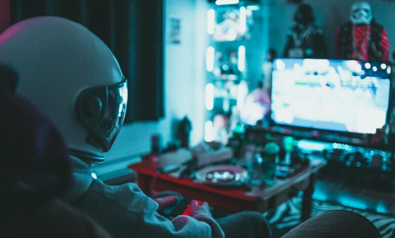 Side view of unrecognizable person in virtual reality helmet sitting on sofa and playing with gamepad in dark room