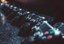 Series of gaming controllers lined up on a reflective surface, dimly lit.