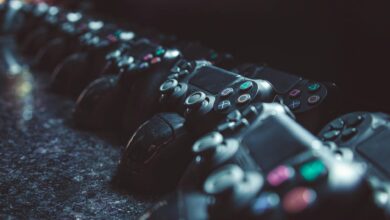 Series of gaming controllers lined up on a reflective surface, dimly lit.
