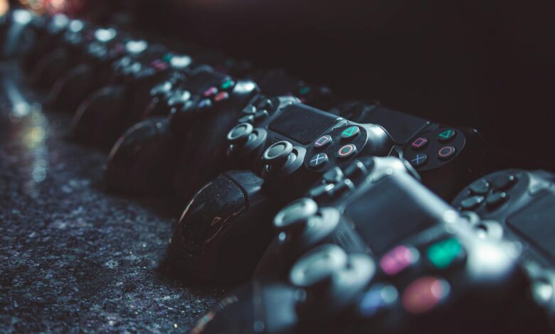 Series of gaming controllers lined up on a reflective surface, dimly lit.
