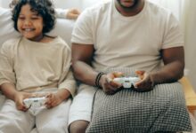 Crop black man and son in sleepwear playing video games on console with joysticks while spending time at home together