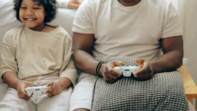 Crop black man and son in sleepwear playing video games on console with joysticks while spending time at home together