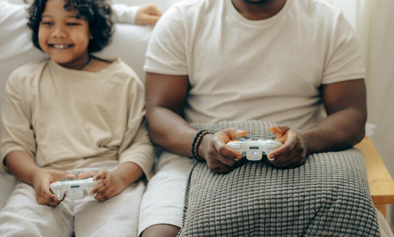 Crop black man and son in sleepwear playing video games on console with joysticks while spending time at home together