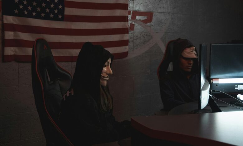 Two masked individuals in a dark room working on computers, symbolizing cyber security threats.
