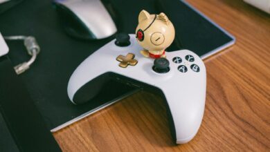 A charming cat figurine sits atop a gaming controller, beside a computer mouse, on a wooden desk.