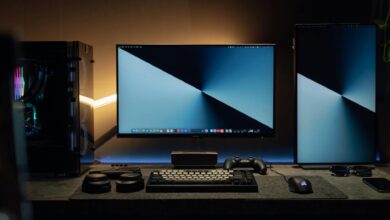 A dimly lit modern computer desk setup featuring dual monitors, a keyboard, and gaming accessories.