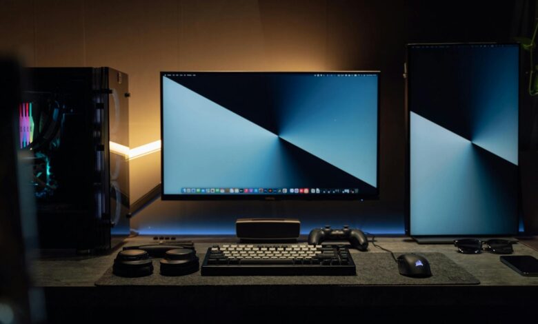 A dimly lit modern computer desk setup featuring dual monitors, a keyboard, and gaming accessories.