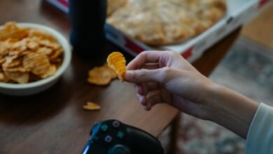 From above of crop unrecognizable man with crispy potato chip and gamepad playing video game in house