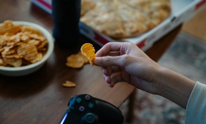 From above of crop unrecognizable man with crispy potato chip and gamepad playing video game in house