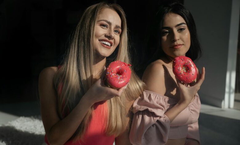 Two women enjoying pink frosted donuts with vibrant sprinkles in a sunlit room.