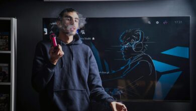 A young man in a hoodie vapes while standing in front of a gaming screen featuring an anime drawing.