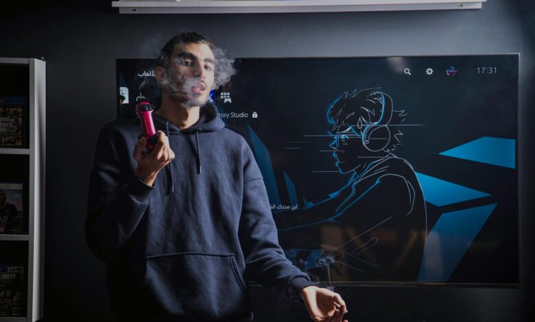 A young man in a hoodie vapes while standing in front of a gaming screen featuring an anime drawing.