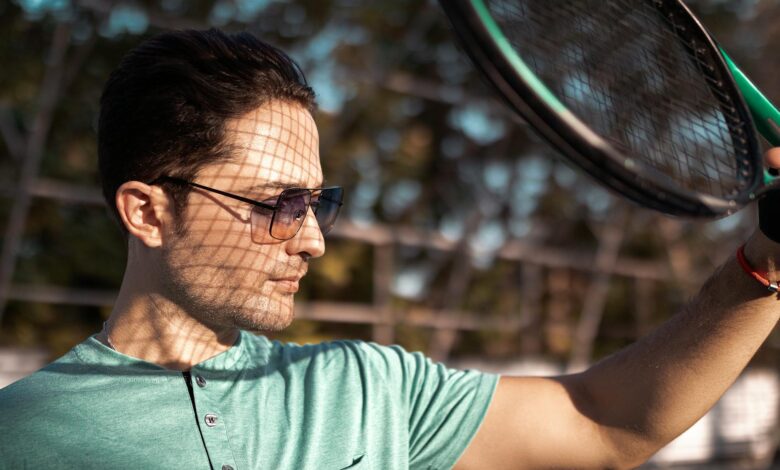Young man in sunglasses holding a tennis racket outdoors on a sunny day.