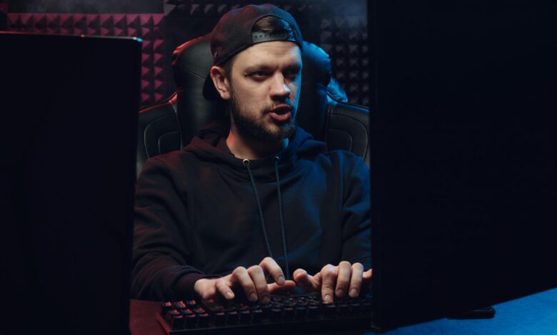Man wearing cap typing on keyboard in a dimly lit room, concentrating on screen.