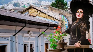 A striking drag queen in elaborate costume poses on a historic Quito balcony with a hand fan.