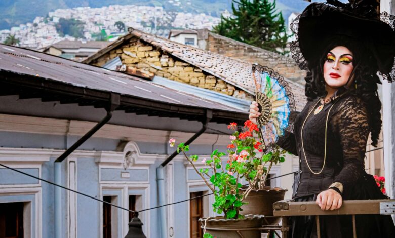 A striking drag queen in elaborate costume poses on a historic Quito balcony with a hand fan.