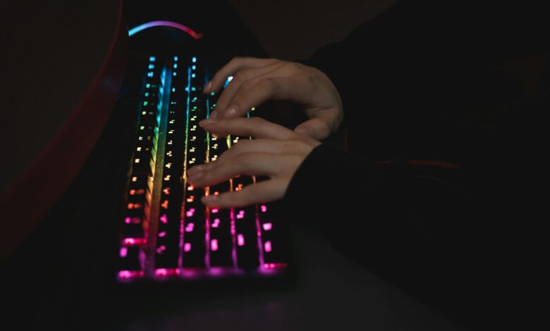 Close-up of hands typing on a colorful RGB backlit mechanical keyboard in a dimly lit environment.