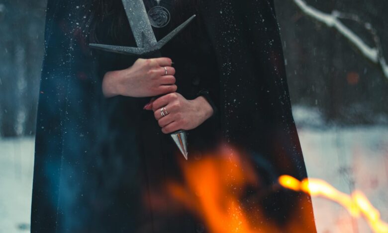 A cloaked figure holds a sword amidst snow and flames, evoking mystery.