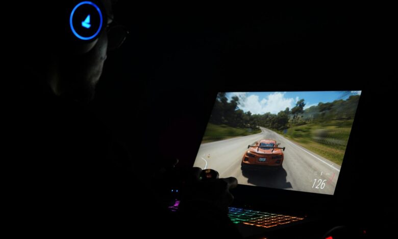 A gamer using a laptop and headphones to play a car racing game in a dark setting.