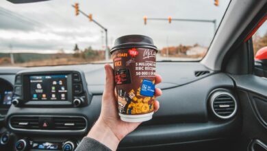 Hand holding coffee cup inside car with intersection view ahead. Captured in overcast weather.