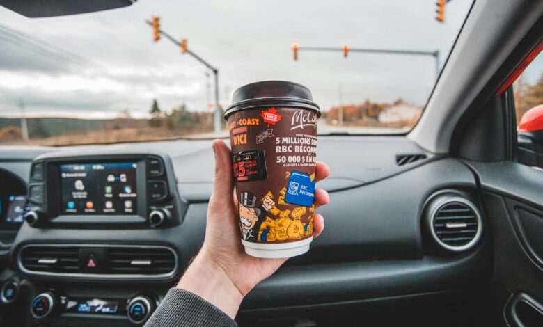 Hand holding coffee cup inside car with intersection view ahead. Captured in overcast weather.