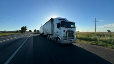 A silver semi truck driving on an open highway under a clear blue sky.