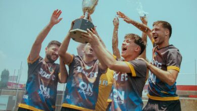 A joyous soccer team celebrates with a trophy outdoors under a clear sky.