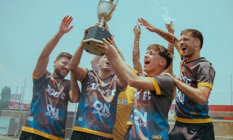 A joyous soccer team celebrates with a trophy outdoors under a clear sky.