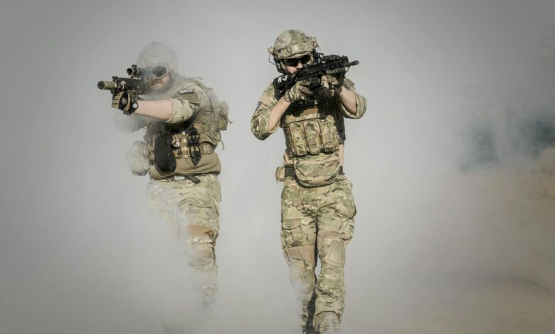 Two soldiers in camo uniforms with weapons advancing through a smoke-covered field, showcasing military action.
