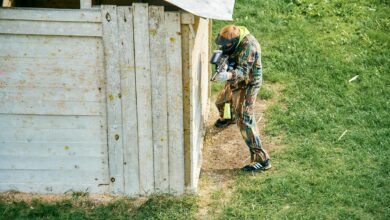 A paintball player taking cover behind a wooden structure on a grassy field during a game.