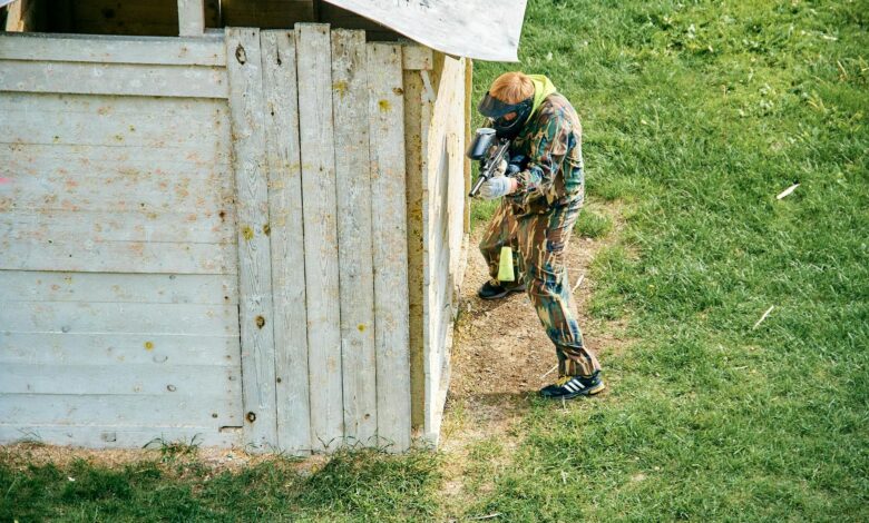 A paintball player taking cover behind a wooden structure on a grassy field during a game.