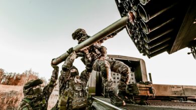 Soldiers in camouflage uniforms loading artillery equipment during a military field operation.