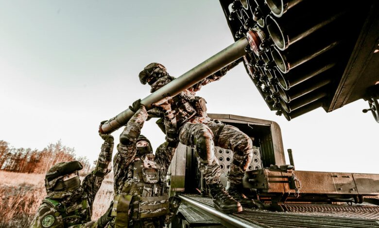 Soldiers in camouflage uniforms loading artillery equipment during a military field operation.
